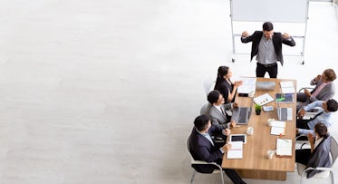 group of sales people gathered around a table looking at a free-standing white board engaged in a meeting lead by a sales leader who uses a structured and simple approach.