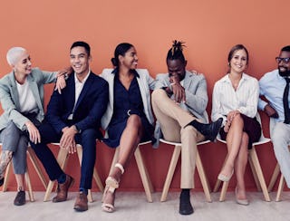 diverse group of young professionals sitting in front of a bold orange wall, looking at the camera representing the diverse, inclusionary, equitable workforce of today.  