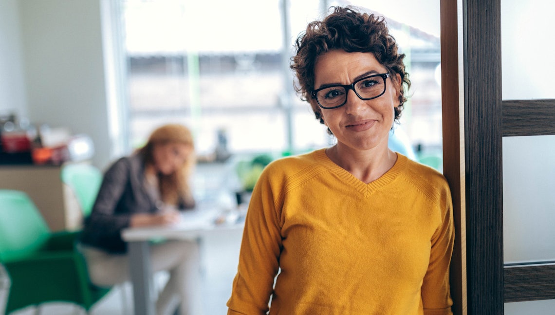 sales manager smiling at camera with a new understanding of what good looks like because of the top performer she just analysed who is sitting in the background,