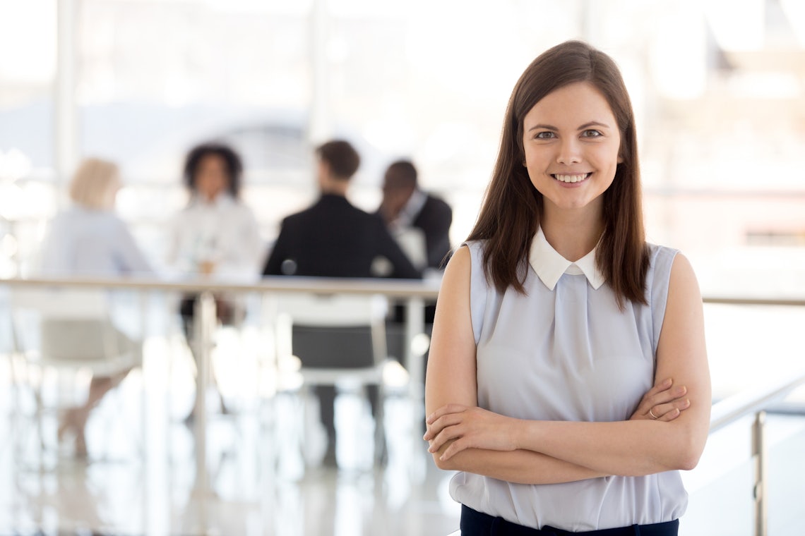 female sales manager looking confidently into the camera while her team works in the background behind her. The team is working hard because she has developed the sales manager traits that build high performing teams.