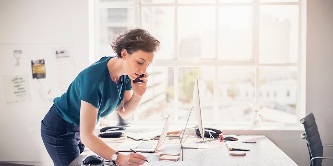 white woman standing over her simplistic desk on her phone on a sunny day connecting with clients to accelerate deals through the pipeline to make her yearly plan.