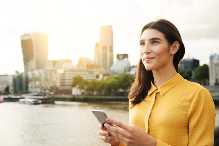 young business woman standing with a phone with a river and city line in the background looking ahead to the future.